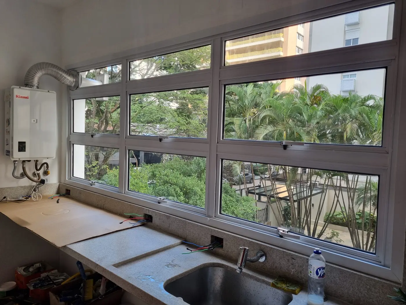 Janela maxim-ar de alumínio com perfil escuro instalada em cozinha com vista para árvores